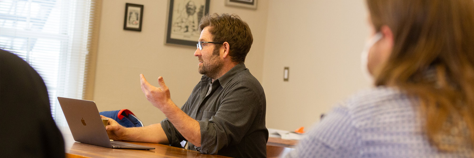 A male teacher, framed by out of focus students on either side of the photograph,  gestures as he makes a point  at the Ostrom Workshop March 2021.