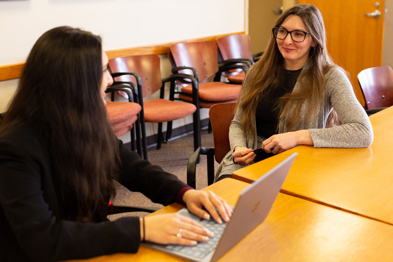 Two young women, one wearing glasses and one typing on a laptop, smile in conversation at the Ostrom Workshop. 