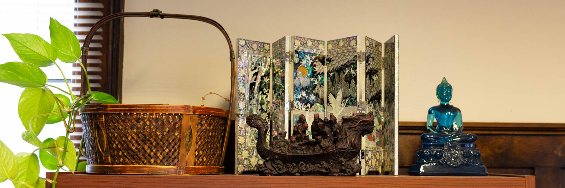 A collection of art and culture-related objects, including a woven basket holding a variegated pothos plant, a decorated miniature Asian room screen, a carved wooden boat, and a blue blown glass statue of the Buddha. 
