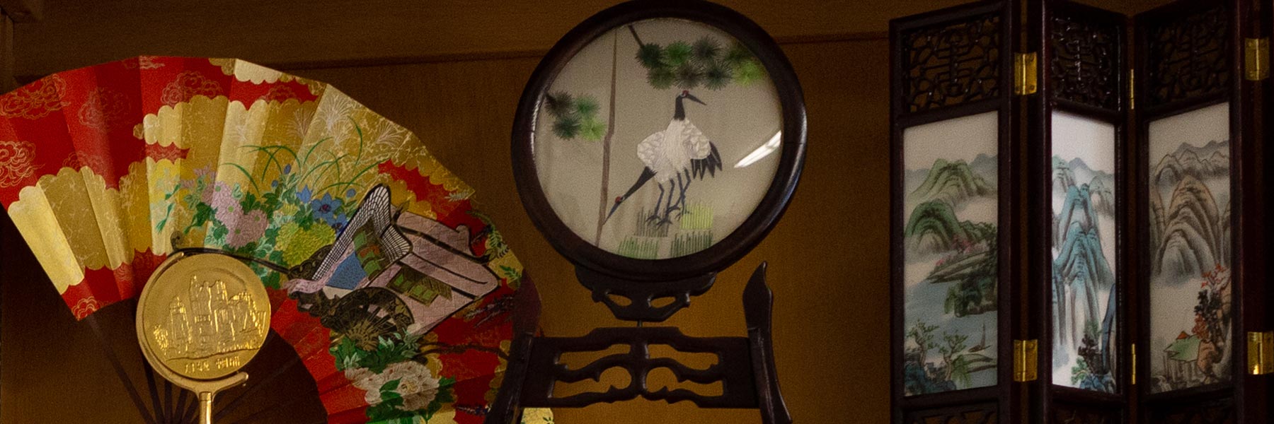 A red and gold Asian fan, a decorative miniature room screen, and a painting of a crane sit together on a shelf. 
